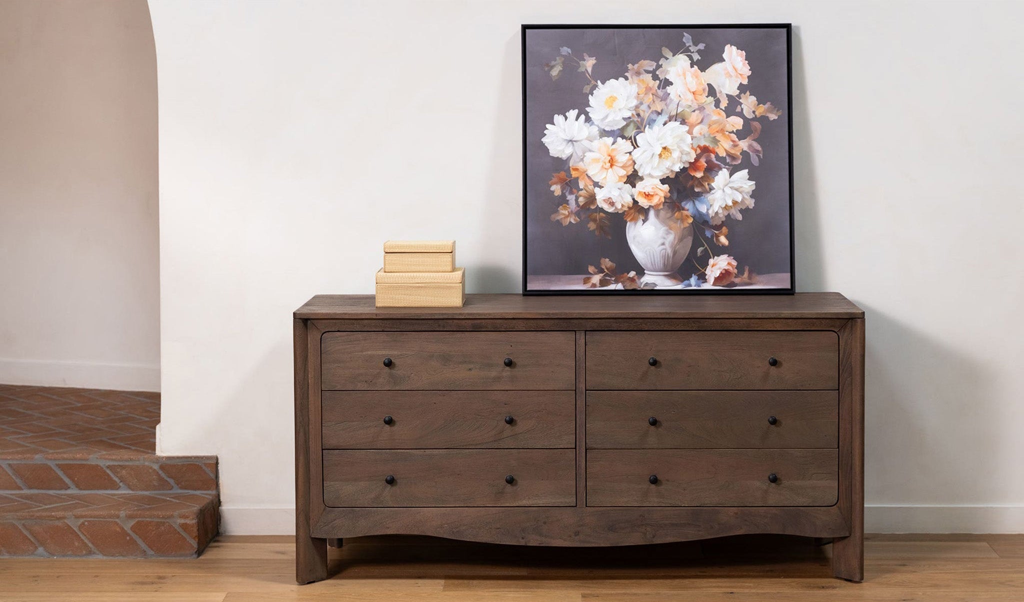 Wooden dresser with a framed floral painting on a white wall.